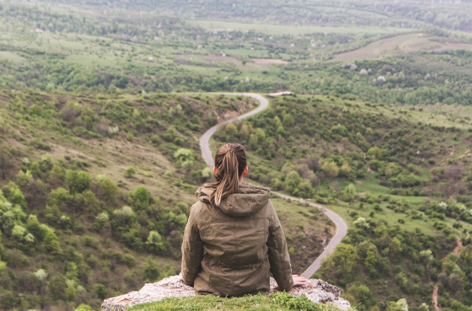 A person seated on a hill overlooking a winding road through a green valley, symbolizing emotional crossroads.