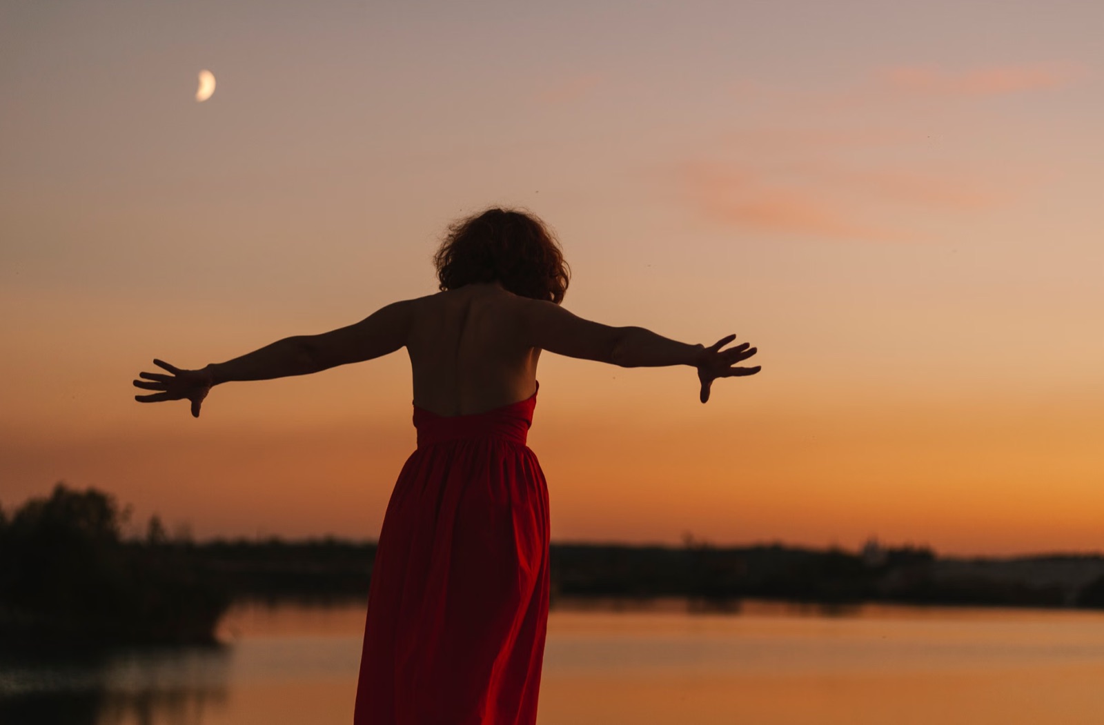 A woman standing calmly by a lake at sunset, symbolizing emotional steadiness and grounded calm.
