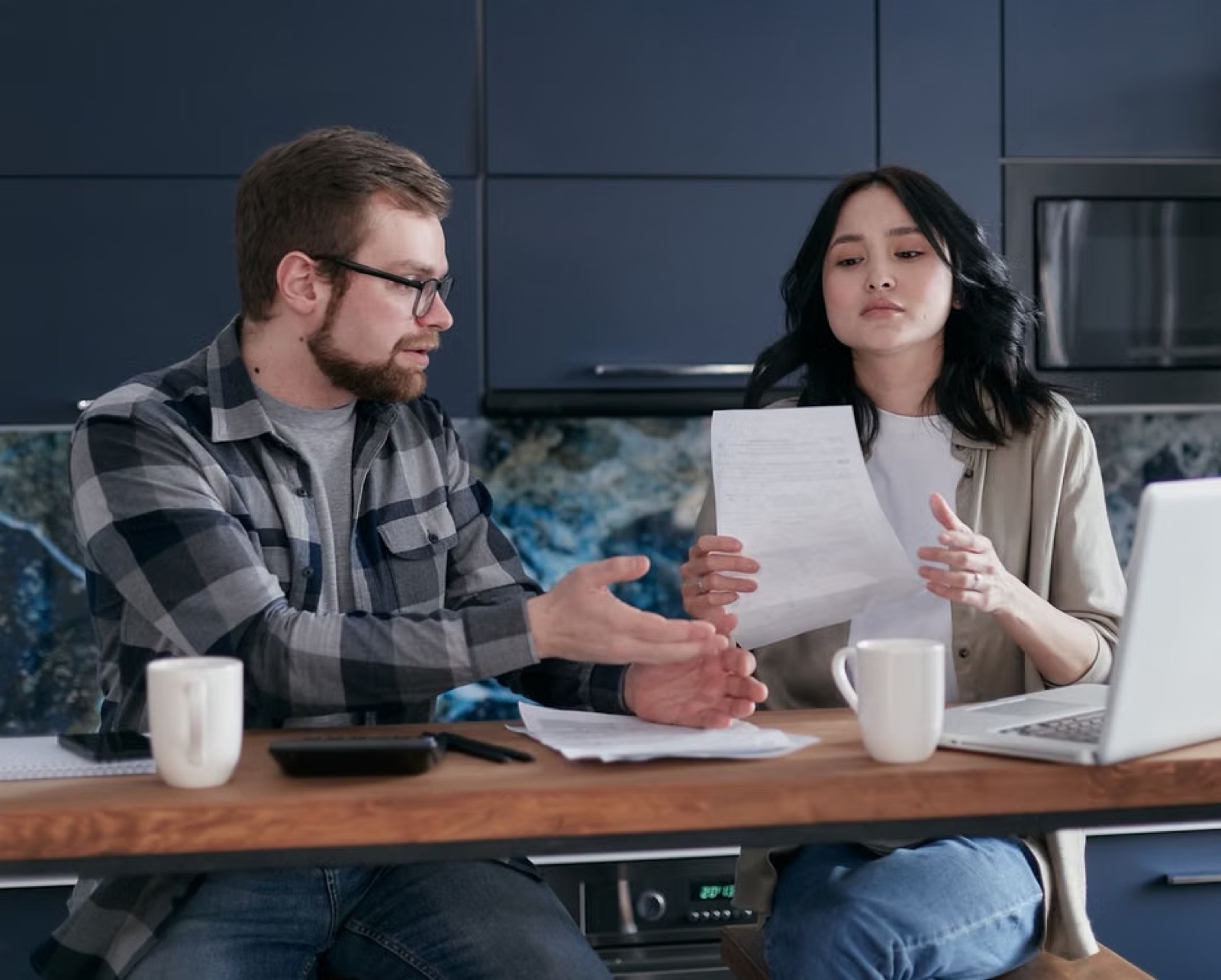 A couple sitting in the same home space with visible tension and practical stress around them.
