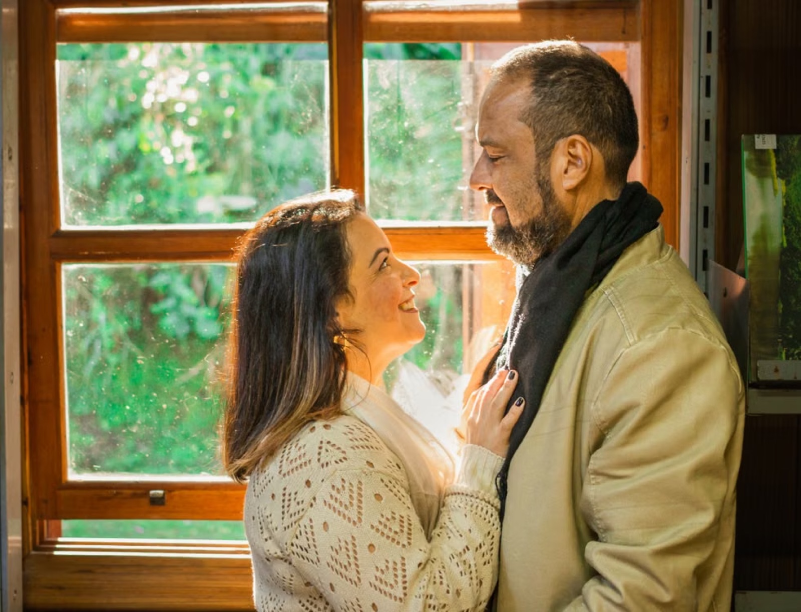 A couple standing close in soft window light, showing intimacy mixed with quiet tension and unspoken feelings.