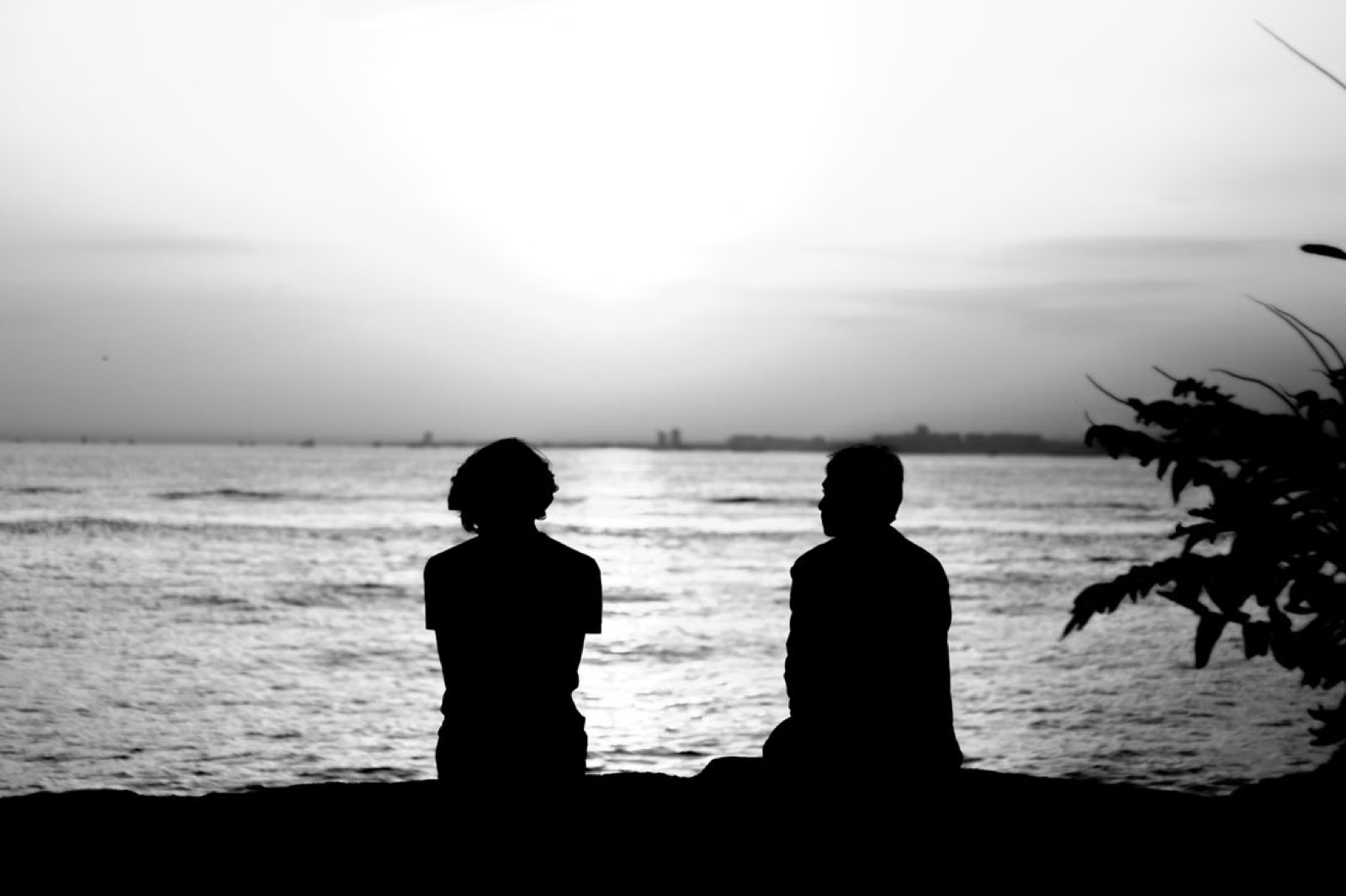 Two people sitting apart by the sea, expressing emotional distance and unspoken relationship tension.