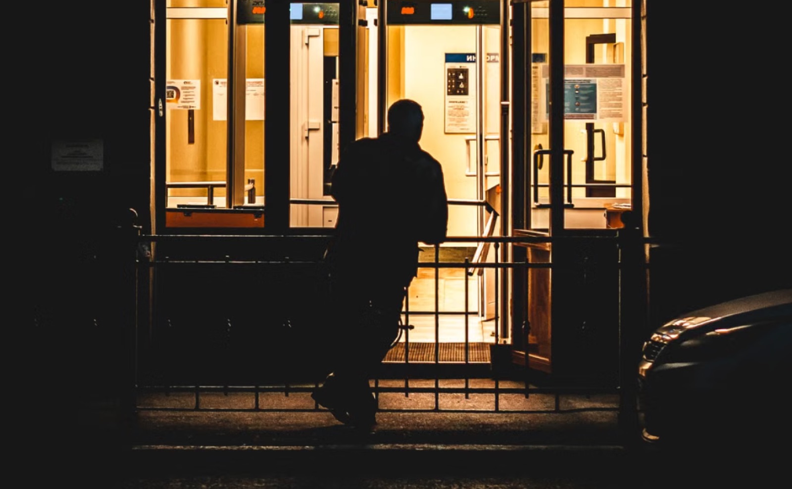 A person hesitating near a warmly lit doorway, suggesting a situation that looks welcoming but does not yet feel settled.