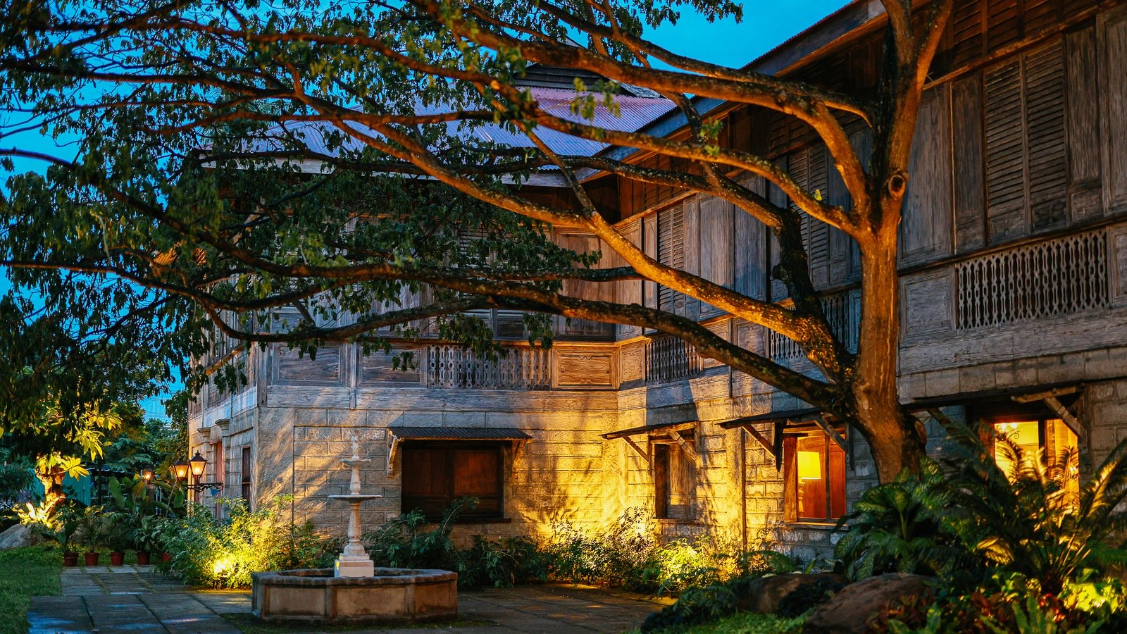 A person standing before a vintage family house with a garden, reflecting the roots and heritage the Ten of Pentacles carries.